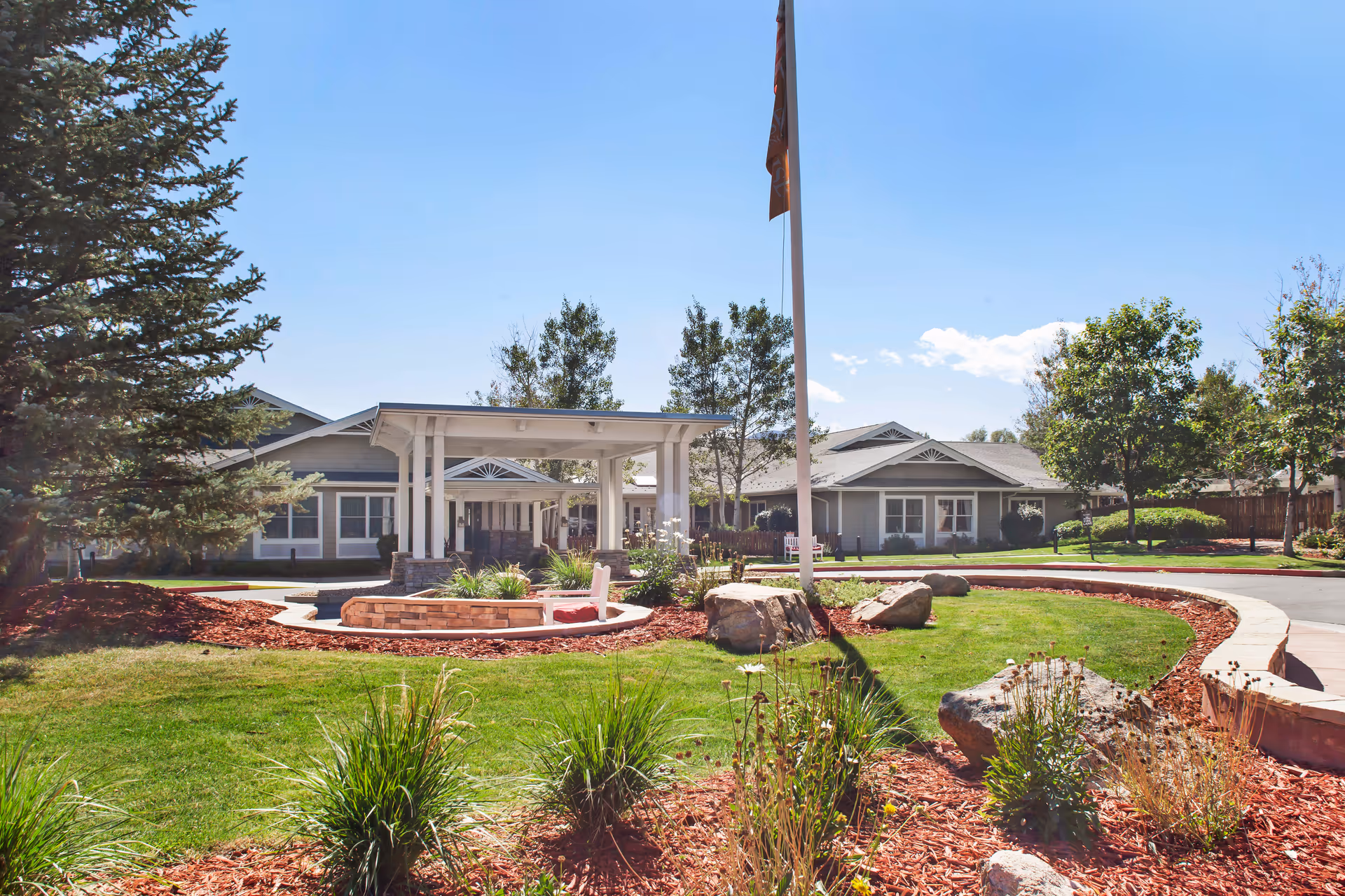 Outdoor view of a senior living facility with a circular driveway, landscaped garden with green grass, shrubs, and rocks. There is a covered entrance with white pillars and a flagpole in the center. The building has a gray exterior with white trim and multiple windows under a clear blue sky.