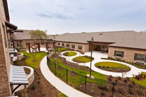Outdoor courtyard area of The Wesleyan Assisted Living and Memory Care facility featuring curved walking paths, green landscaped areas with grass and small trees, black metal fencing, and a covered seating area with wooden pergola. The courtyard is surrounded by a single-story brick building under a cloudy sky.