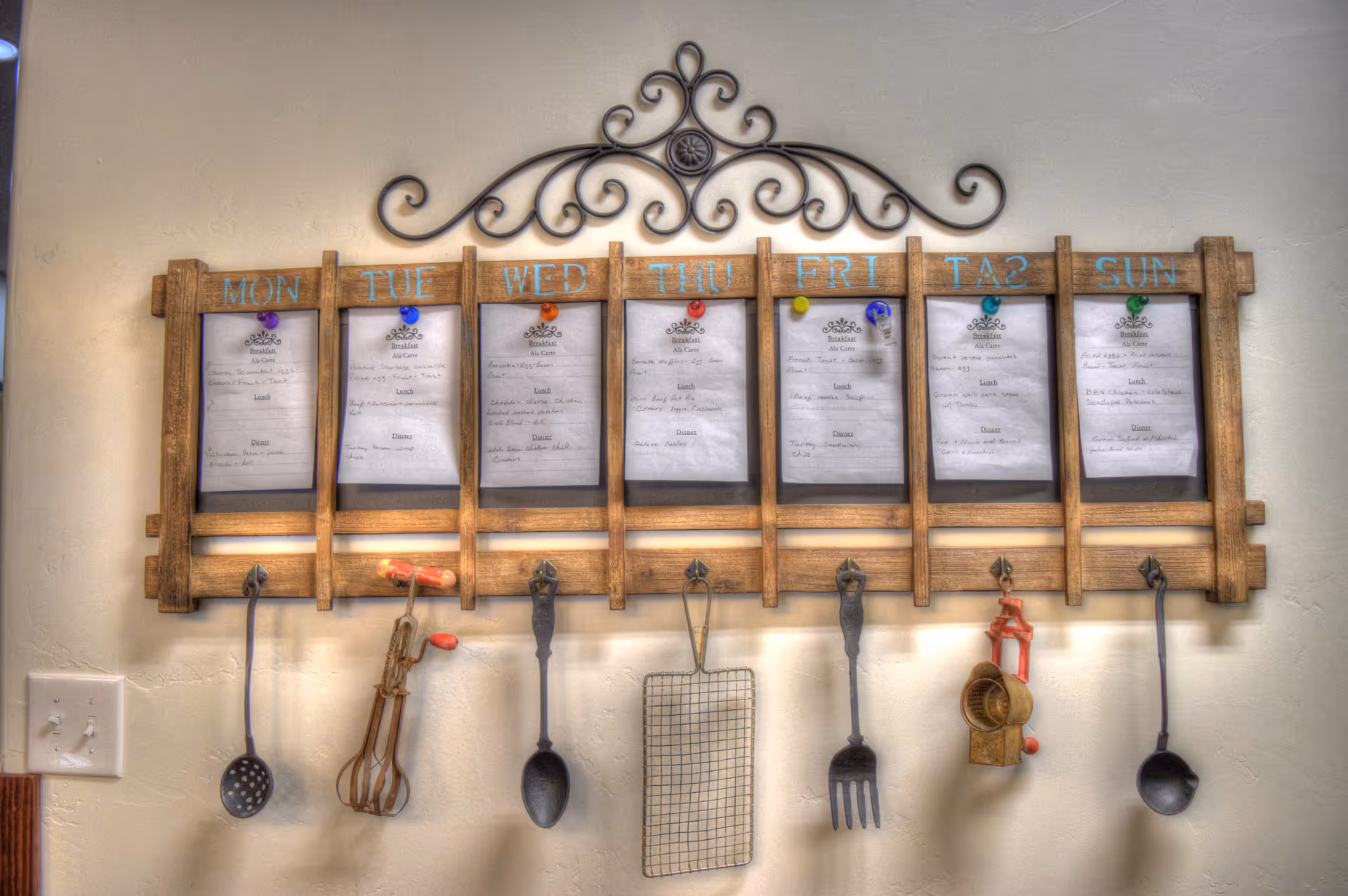 A wooden weekly menu board mounted on a wall with days of the week labeled from Monday to Sunday. Each day has a pinned menu sheet listing breakfast, lunch, and dinner options. Below the menu are hanging vintage kitchen utensils including ladles, a hand mixer, a large spoon, a wire rack, a fork, and a manual food grinder. The board has decorative metal scrollwork at the top.
