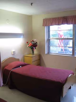 Single bed with maroon cover, bedside table, flowers, and window in a small room