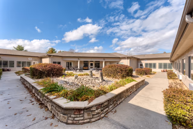 Outdoor courtyard area at Cedarhurst Senior Living of Springfield, MO, featuring a stone planter with bushes and plants, surrounded by a concrete walkway. The building surrounds the courtyard with large windows and a covered patio area with tables and chairs under a partly cloudy blue sky.