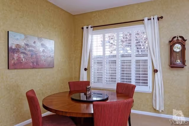 Small dining room with a round wooden table surrounded by four red upholstered chairs, a window with white shutters and curtains, wall art, and a hanging clock.