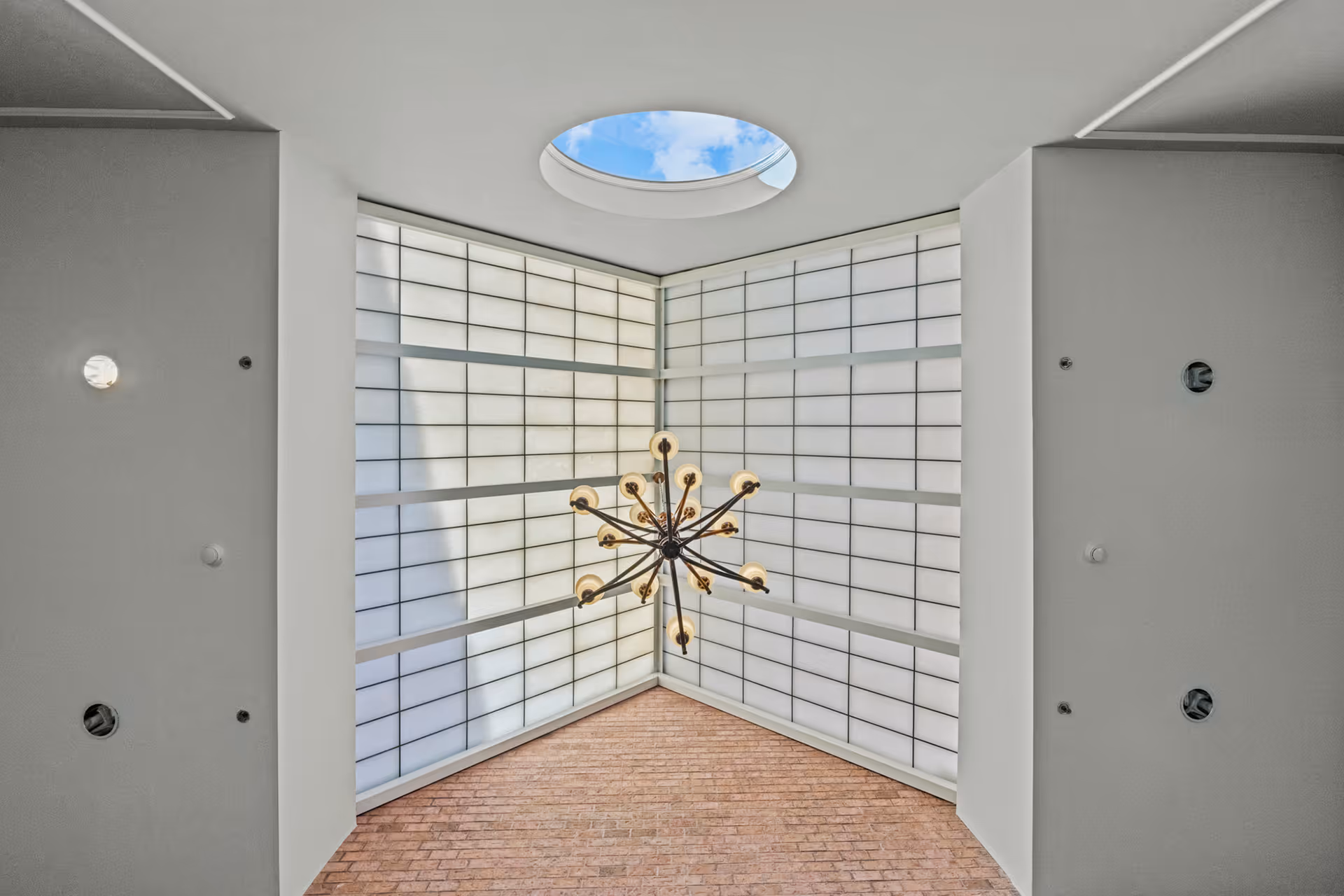 View of a modern ceiling with a large grid-patterned skylight and a circular opening showing blue sky and clouds. A contemporary chandelier with multiple light bulbs hangs from the ceiling above a brick wall.