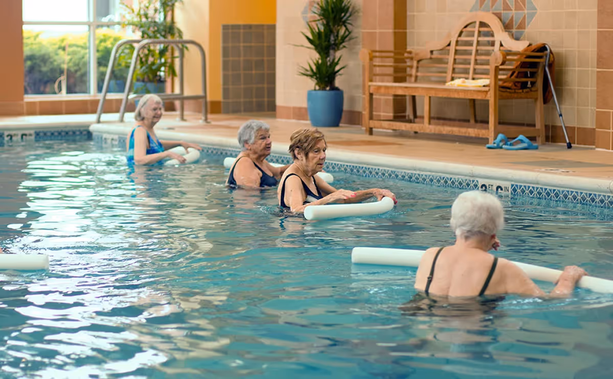 Four elderly women participating in a water exercise class in an indoor swimming pool, each holding a white pool noodle. The pool area has large windows, a wooden bench, potted plants, and a tiled wall.