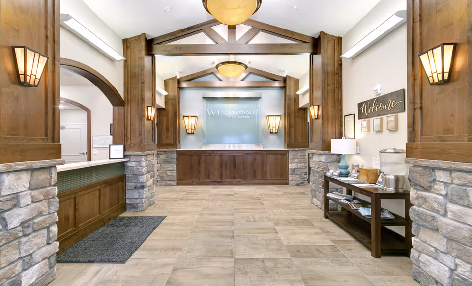 Reception area of Windsong at Northridge featuring a wooden front desk with stone accents, warm wall sconces, a welcome sign, and a table with a lamp and water dispenser.