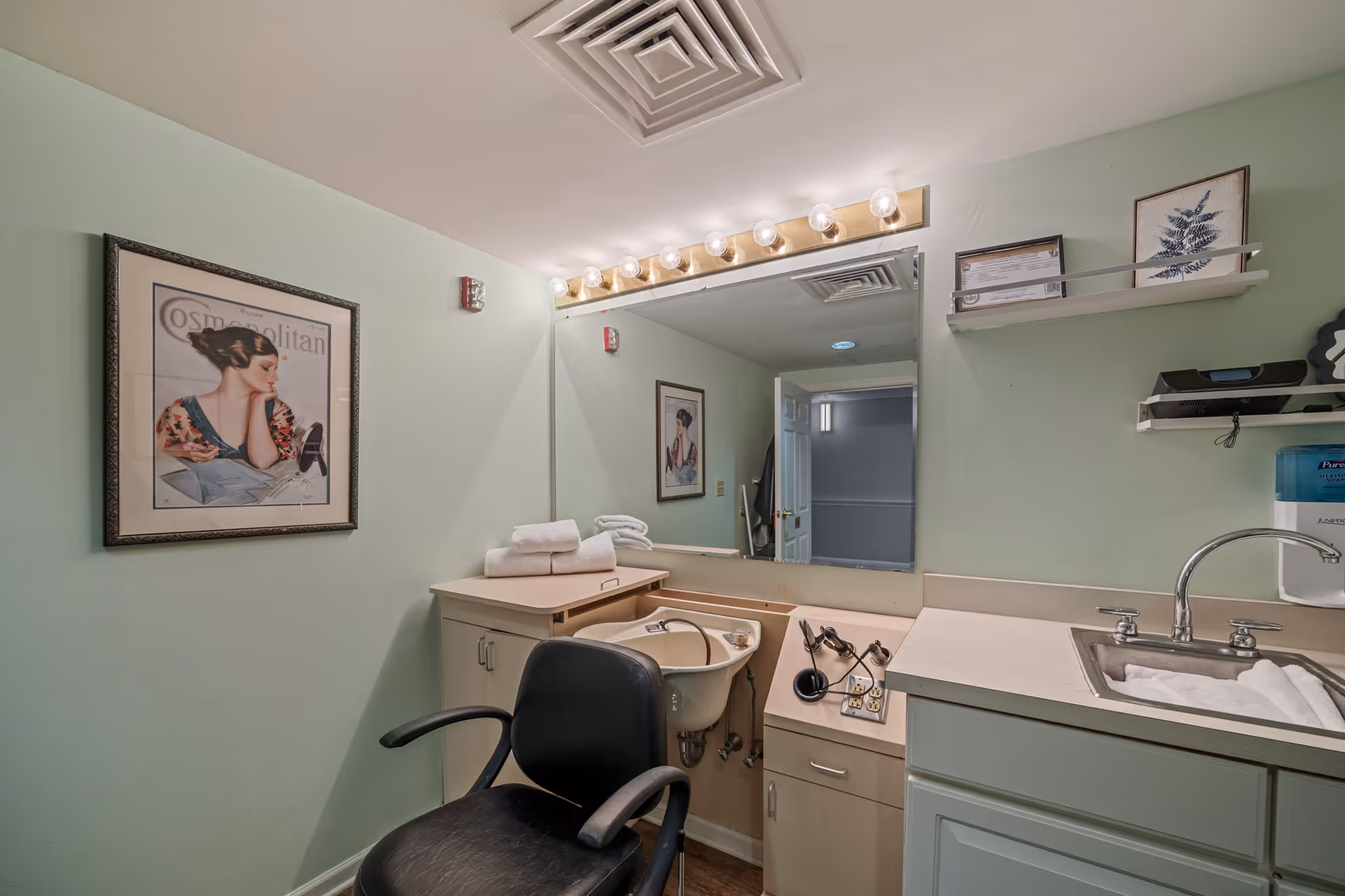 Hair-washing salon station with a black styling chair, shampoo sink, mirror with vanity lights, countertop and framed art on mint-green walls.