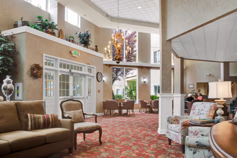 Sunlit, high-ceilinged senior living lobby with multiple seating areas, a chandelier, patterned carpet, and large windows.