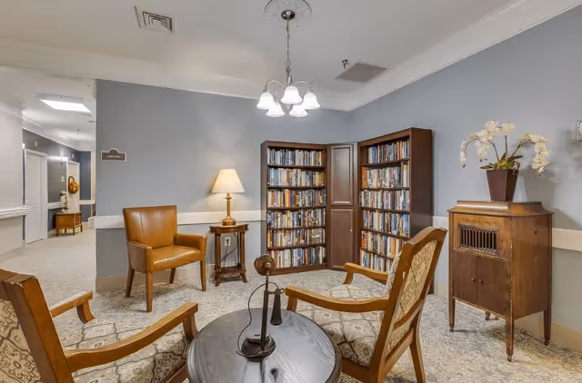 A cozy reading nook in a senior living facility featuring a round table with an antique telephone, surrounded by four wooden armchairs with patterned cushions. Behind the chairs are two tall bookshelves filled with books, a small wooden cabinet with a potted orchid on top, and a side table with a lamp. The walls are painted light gray and the floor is carpeted. A hallway is visible in the background.