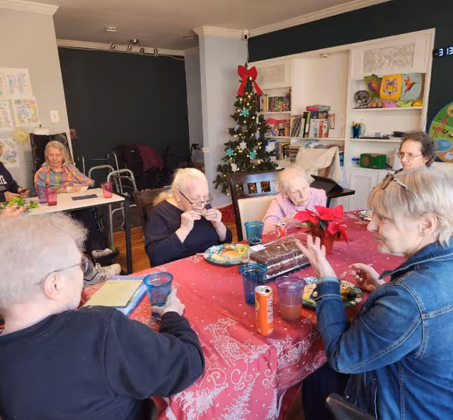A group of elderly people sitting around a table covered with a red holiday-themed tablecloth, eating and socializing. There is a decorated Christmas tree with a red bow on top in the background, along with bookshelves and holiday decorations. The setting appears to be a cozy common area in a senior living facility.
