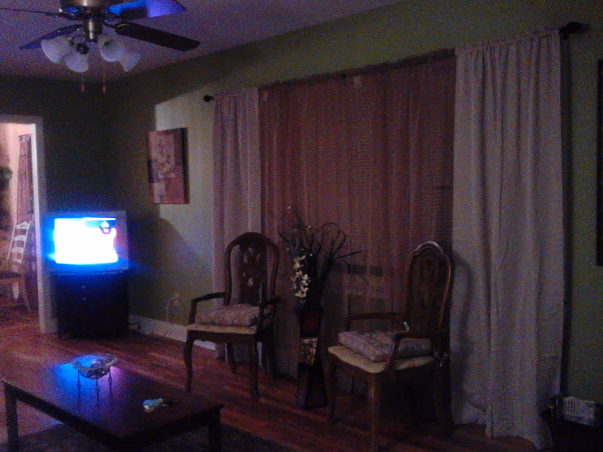 A dimly lit living room with two wooden chairs with cushions placed against a wall with large windows covered by curtains. A small TV is on a stand in the corner, and a coffee table with a decorative bowl and a remote control is in the foreground. A ceiling fan with lights is visible above.