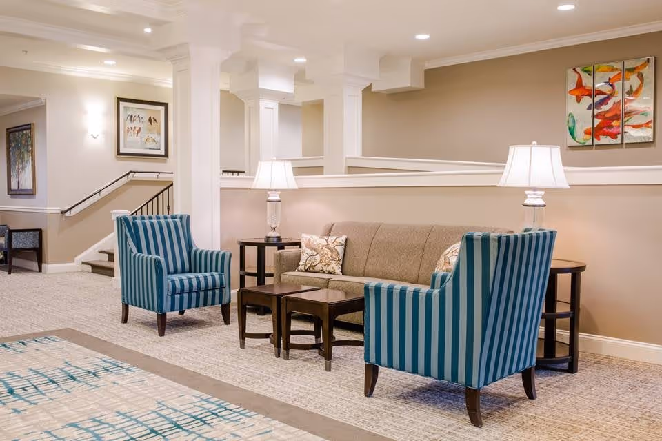 A cozy seating area in a senior living facility with a beige sofa, two blue and green striped armchairs, two side tables with lamps, and colorful artwork on the walls. The space is well-lit with recessed ceiling lights and has a carpeted floor.