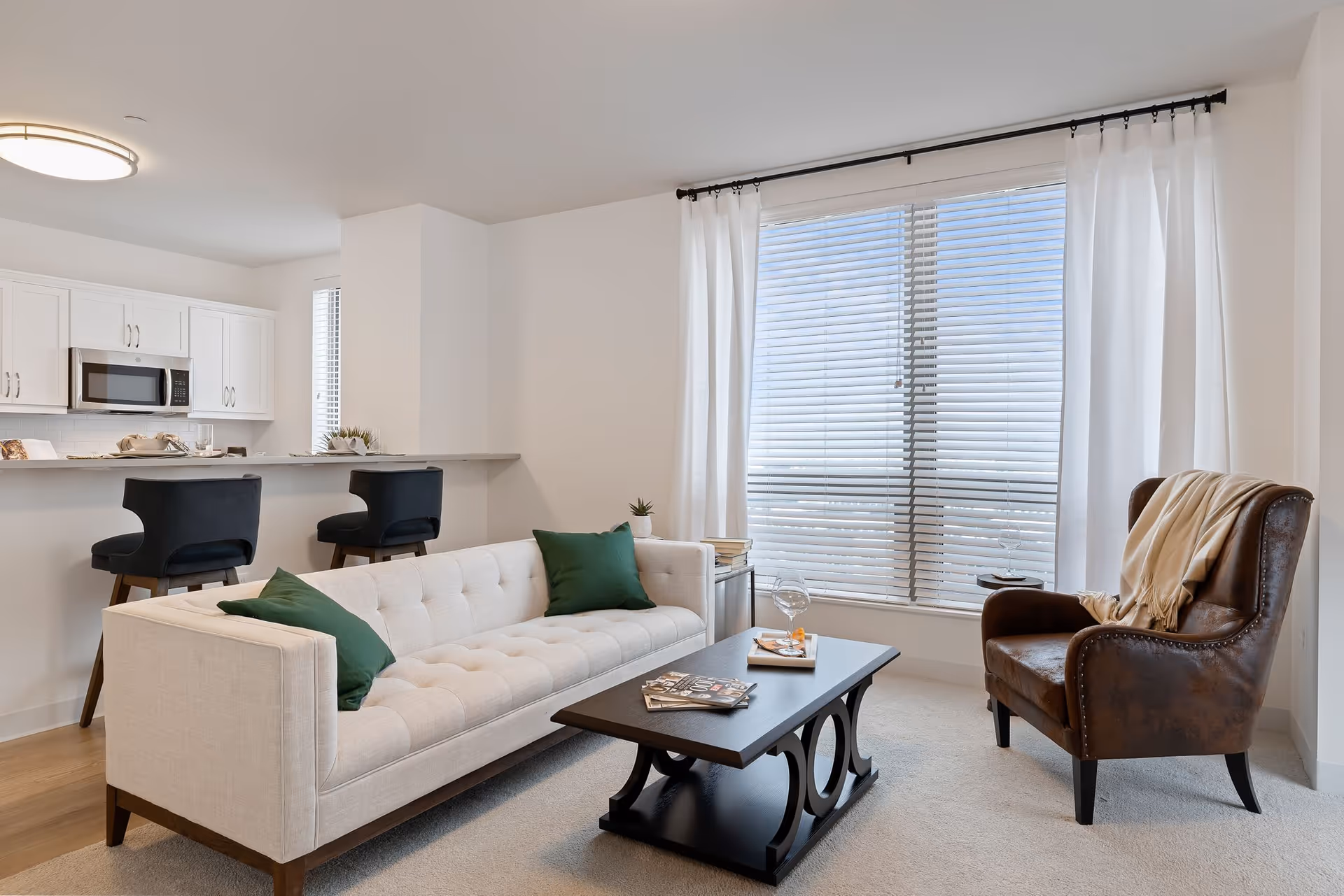 A bright and modern living room with a beige tufted sofa adorned with green pillows, a dark wooden coffee table with magazines and a glass of wine, a brown leather armchair with a beige throw draped over it, and large windows with white curtains and blinds letting in natural light. In the background, a kitchen area with white cabinets, a microwave, and two black bar stools at a counter is visible.