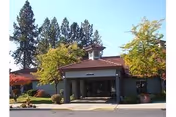 Front exterior view of Fairwood Retirement Village & Assisted Living building with a covered entrance, surrounded by trees and landscaping under a clear blue sky.