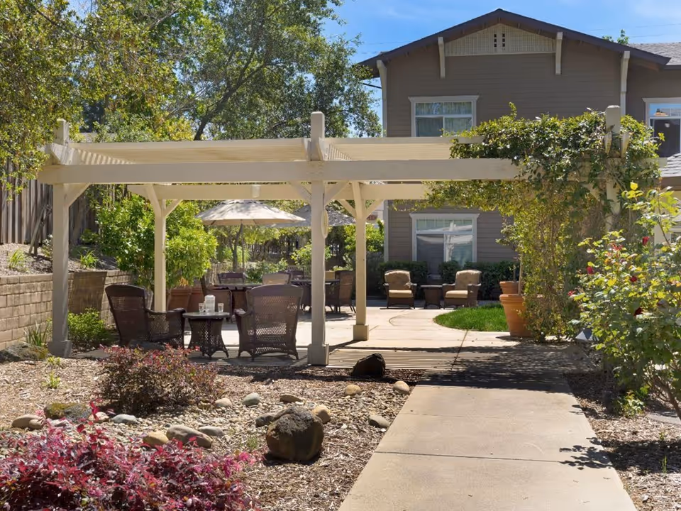 Outdoor patio area with a white pergola, several wicker chairs and tables, an umbrella providing shade, surrounded by plants, bushes, and trees, with a two-story building in the background under a clear blue sky.