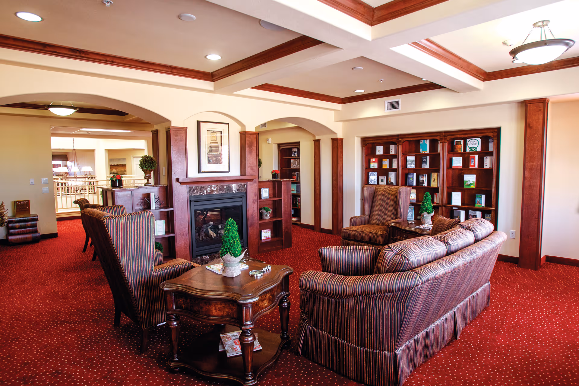 A cozy living room area with red carpet and wooden furniture including a striped sofa and armchairs arranged around a wooden coffee table. There is a fireplace with a framed picture above it and built-in bookshelves filled with books and decorative items. The ceiling has recessed lighting and wooden beams.