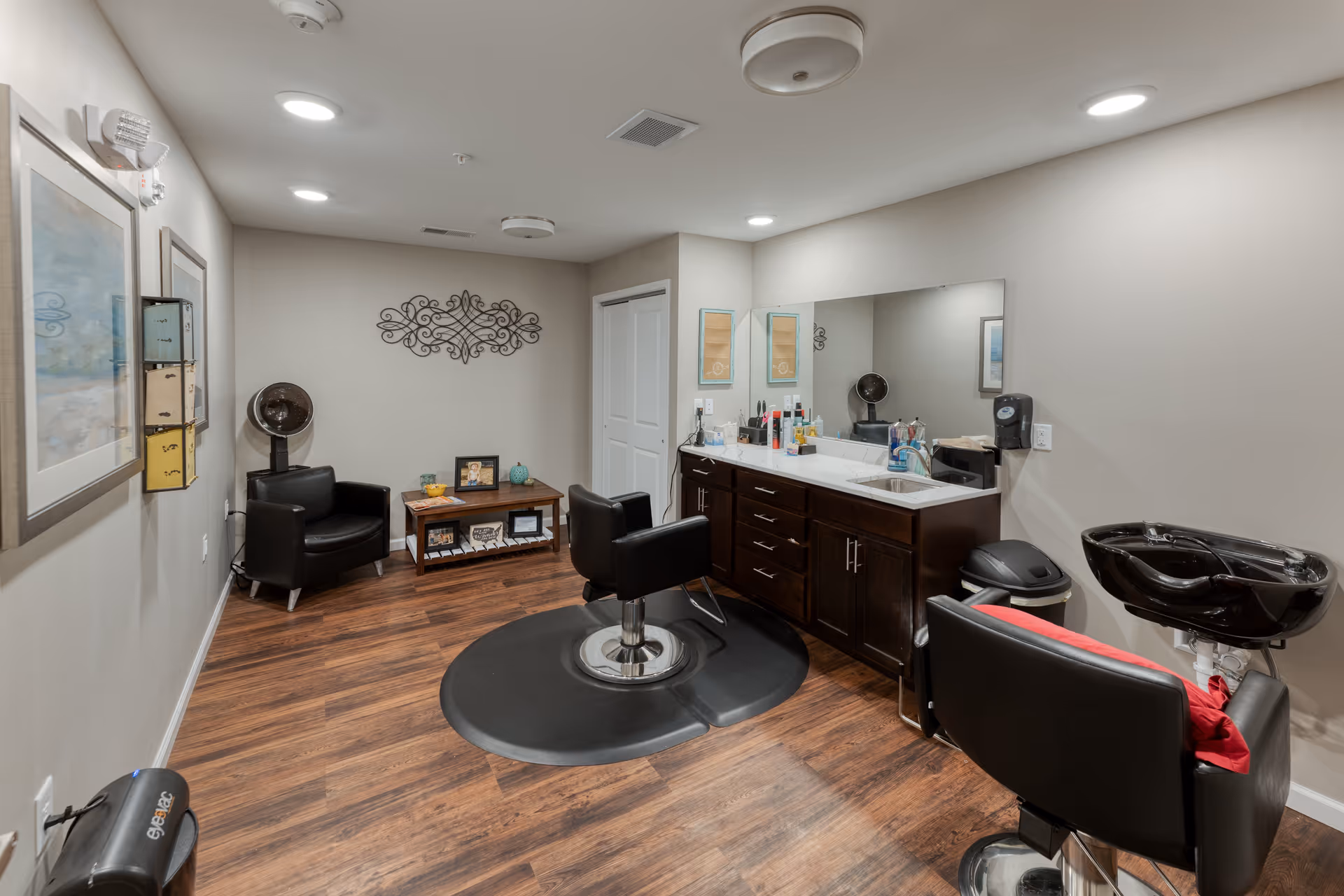 A well-lit salon room with wooden flooring featuring a black salon chair on a protective mat in front of a large mirror and a countertop with various hair care products. There is a black shampoo basin chair with a red cushion, a black armchair, a small wooden table with decorative items, and wall art. The walls are painted light gray.