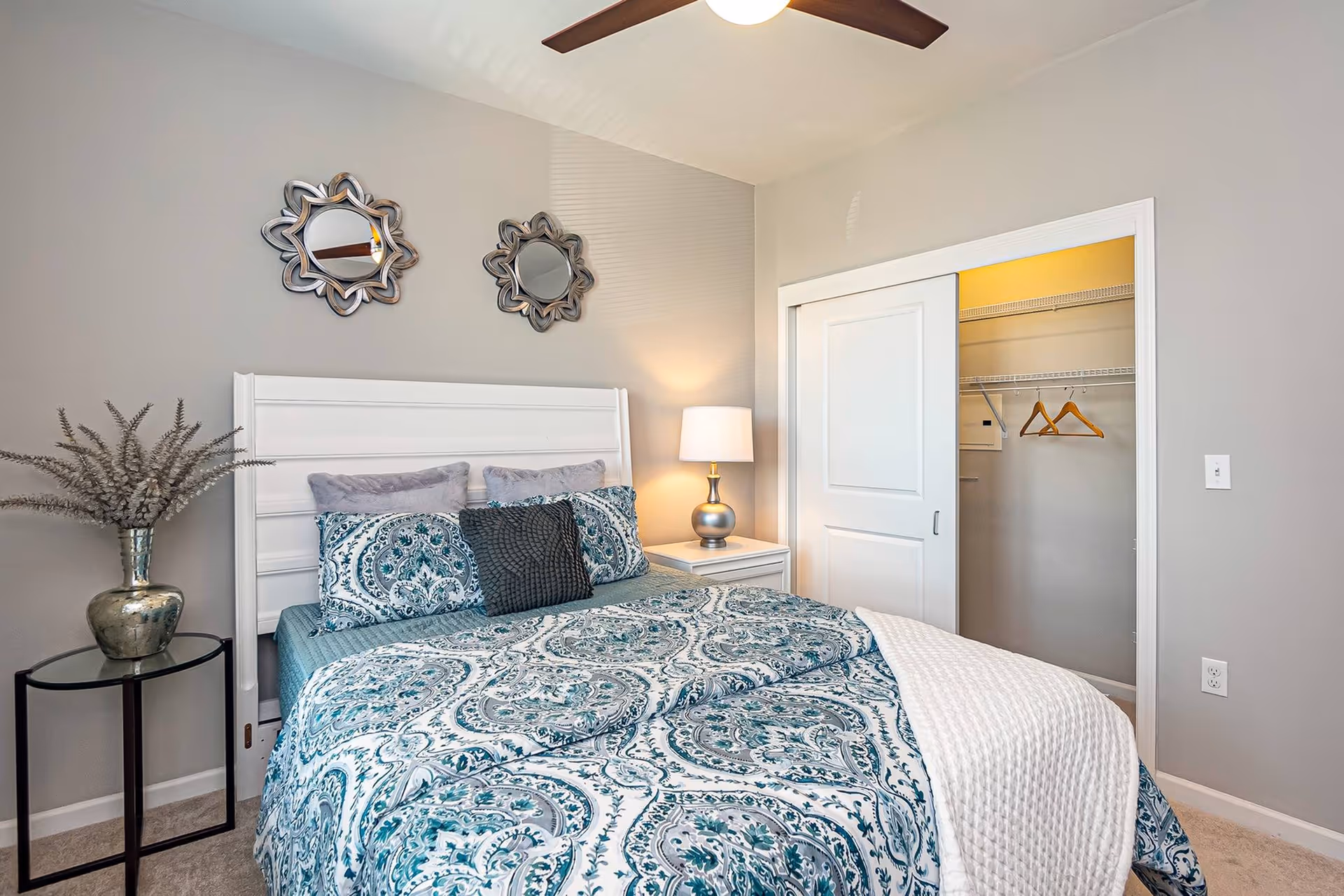 A neatly made bed with a white headboard and blue patterned bedding in a bedroom. There are two decorative mirrors on the wall above the bed, a bedside table with a lamp, and a small round table with a vase holding dried plants. A closet with sliding doors is partially open, showing hangers inside.