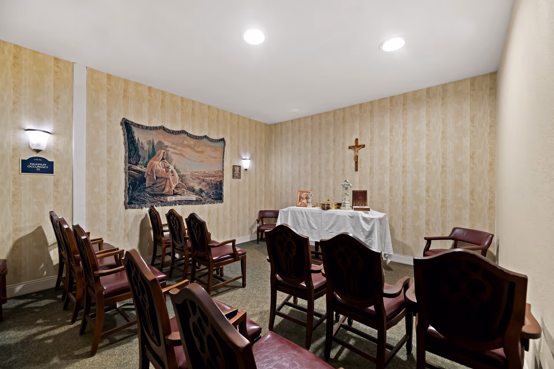 A small chapel room with wooden chairs arranged in rows facing a table covered with a white cloth. On the table are religious items including a cross, a framed picture, and a book. The walls have beige striped wallpaper, a tapestry depicting a religious scene, and a crucifix hanging above the table. Two wall sconces provide lighting.
