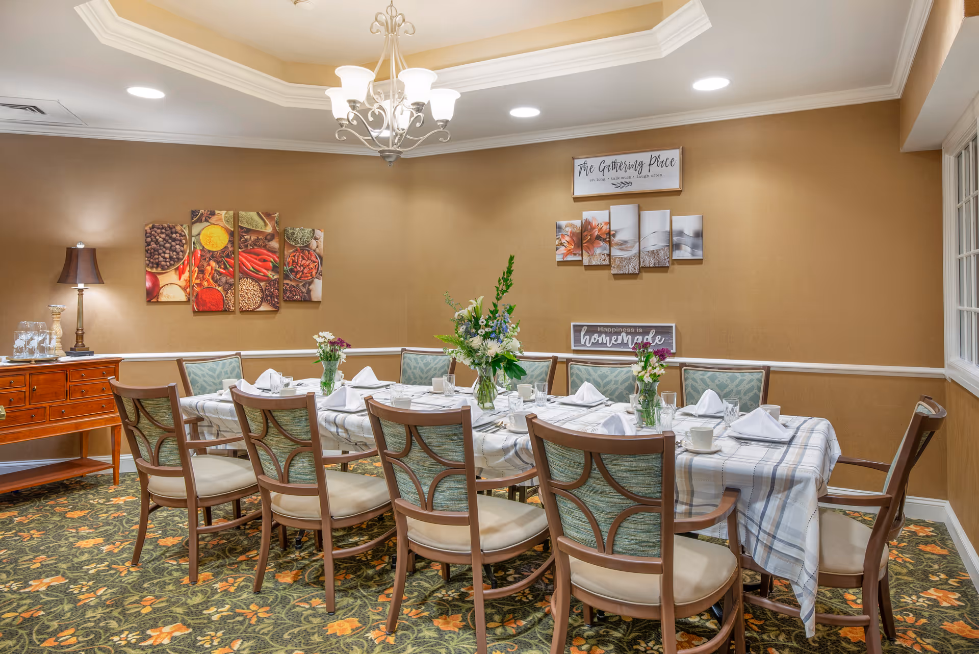 A dining room with a long table covered with a checkered tablecloth, set with white napkins, plates, glasses, and floral centerpieces. The room has a patterned carpet, beige walls with framed artwork and signs, a chandelier, and a wooden sideboard with a lamp and glassware.