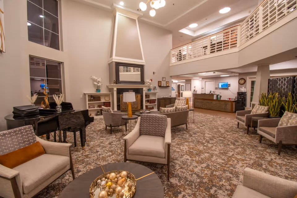 A spacious and well-lit living room area in a senior living facility featuring comfortable armchairs arranged around small tables, a grand piano, a large fireplace with a mirror above it, and a reception desk in the background. The room has high ceilings with a balcony overlooking the space and decorative plants along one side.