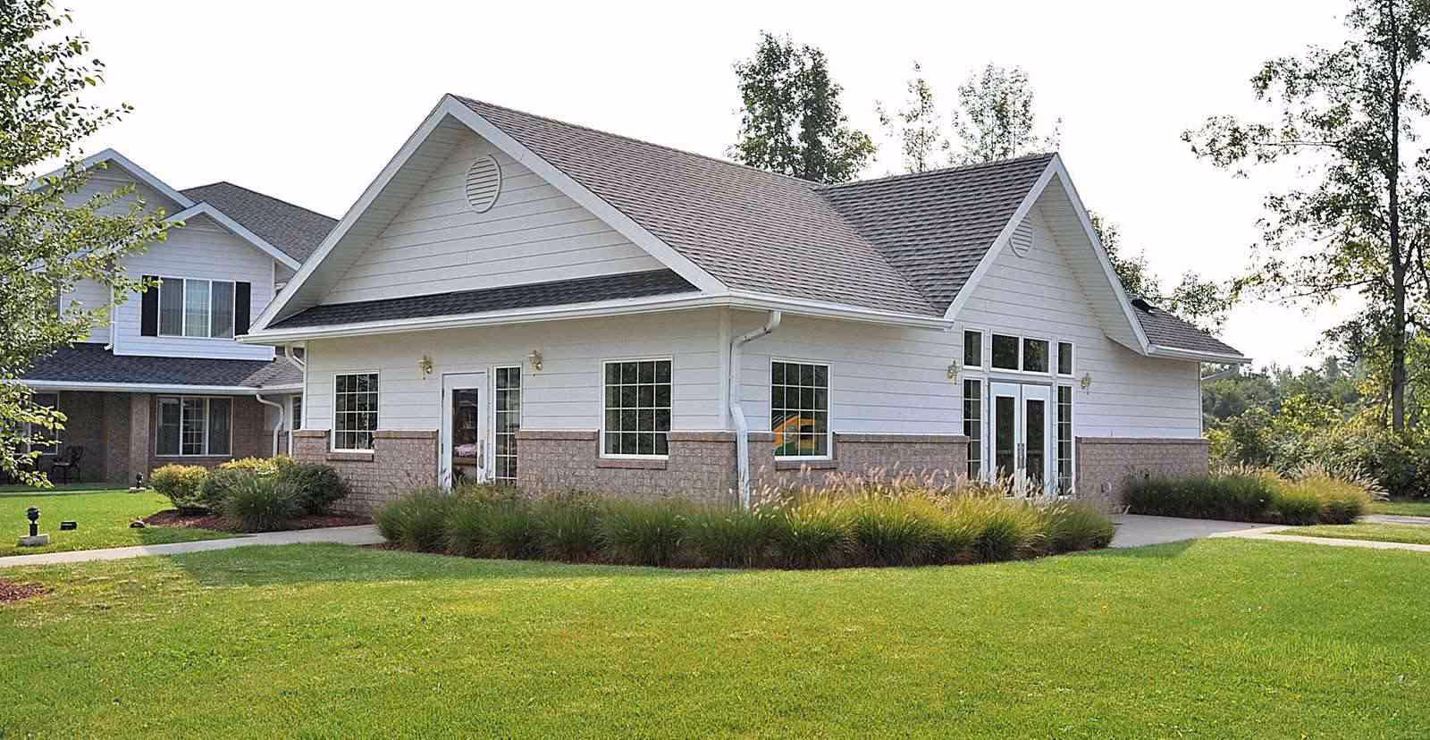 Exterior view of a single-story building with white siding and brick accents, surrounded by green grass and landscaping. The building has multiple windows and a gabled roof. Trees and another building are visible in the background.
