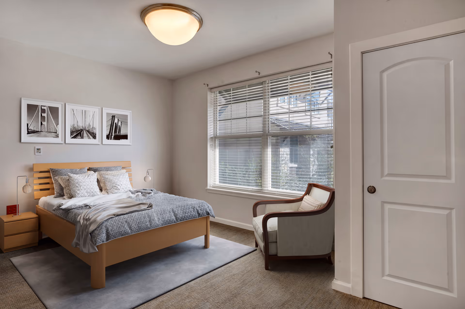 A bright bedroom featuring a wooden bed with gray bedding, nightstands and lamps, framed black-and-white photos above the bed, a large window with blinds, and an upholstered armchair.