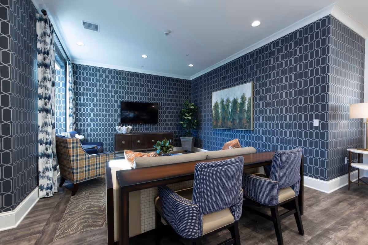 A modern living room with dark geometric patterned wallpaper, a wall-mounted flat screen TV, a wooden console table, a beige sofa with orange and patterned throw pillows, two blue patterned armchairs, a plaid armchair near a window with patterned curtains, a potted plant in the corner, a framed painting of trees on the wall, and a floor lamp beside a small side table.