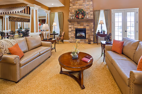 A cozy living room with two tan leather sofas facing each other, each with orange throw pillows. A round wooden coffee table with a small plant and books sits between the sofas. The room features a stone fireplace decorated with a wreath, flanked by windows with curtains and lamps on side tables. In the background, there is a dining area with tables and chairs, and a striped awning over part of the space.