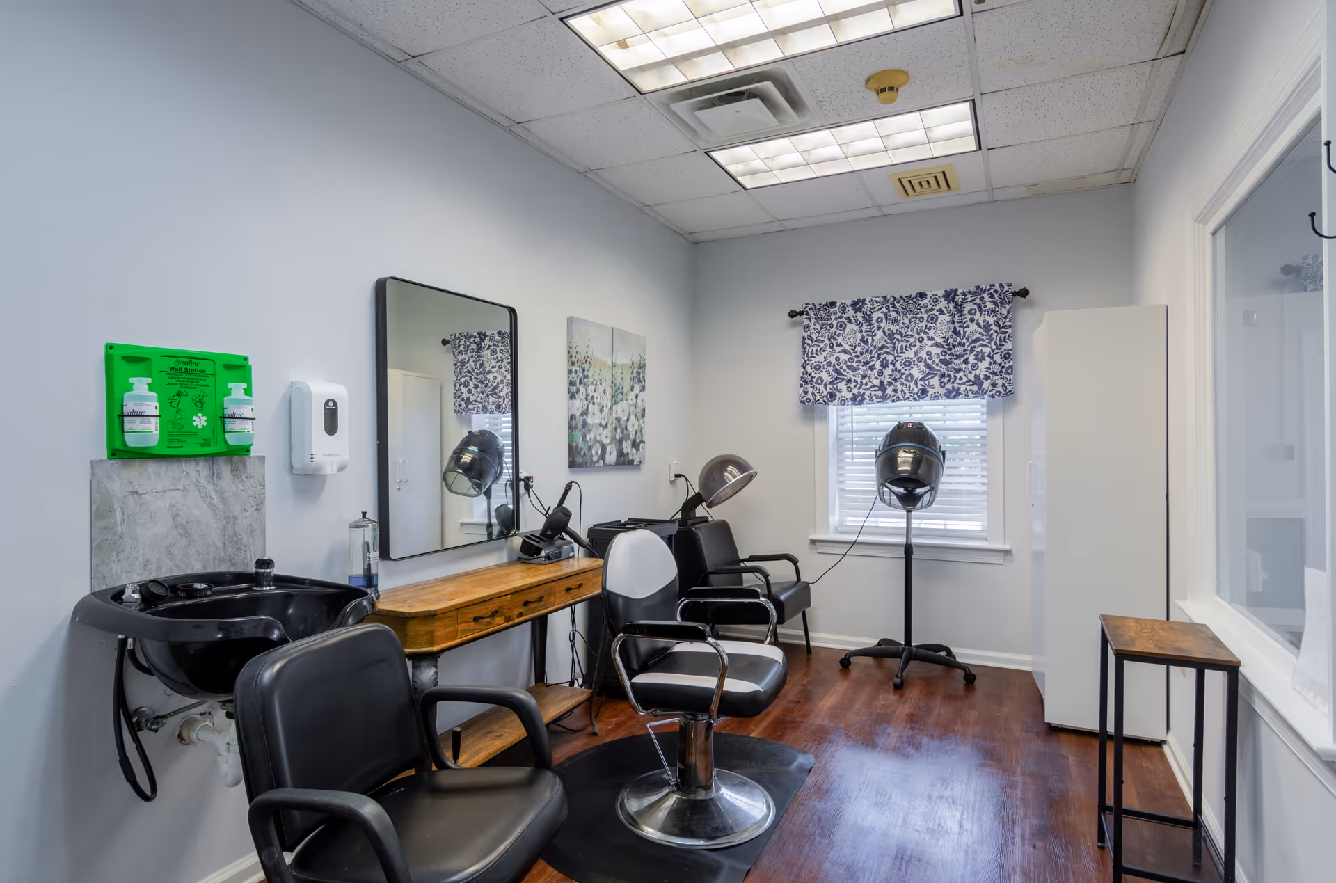 Interior of a small salon room with two black salon chairs, a black hair washing sink, a wooden counter with a mirror above it, hair drying equipment, a window with blue and white floral curtains, and a wooden floor.