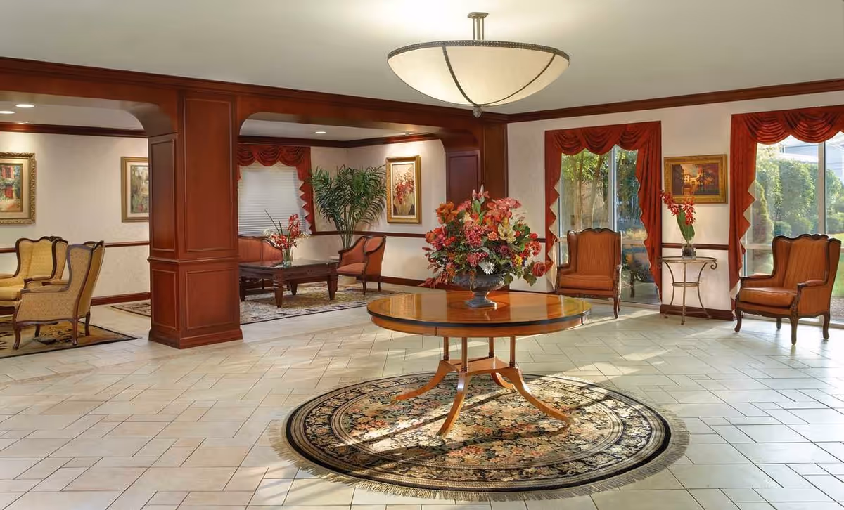 A spacious, well-lit common area with a round wooden table in the center adorned with a large floral arrangement. The room features tiled flooring with a circular rug under the table, wooden columns, and several seating areas with upholstered chairs and small tables. Large windows with red curtains allow natural light to brighten the space, and framed artwork decorates the walls.