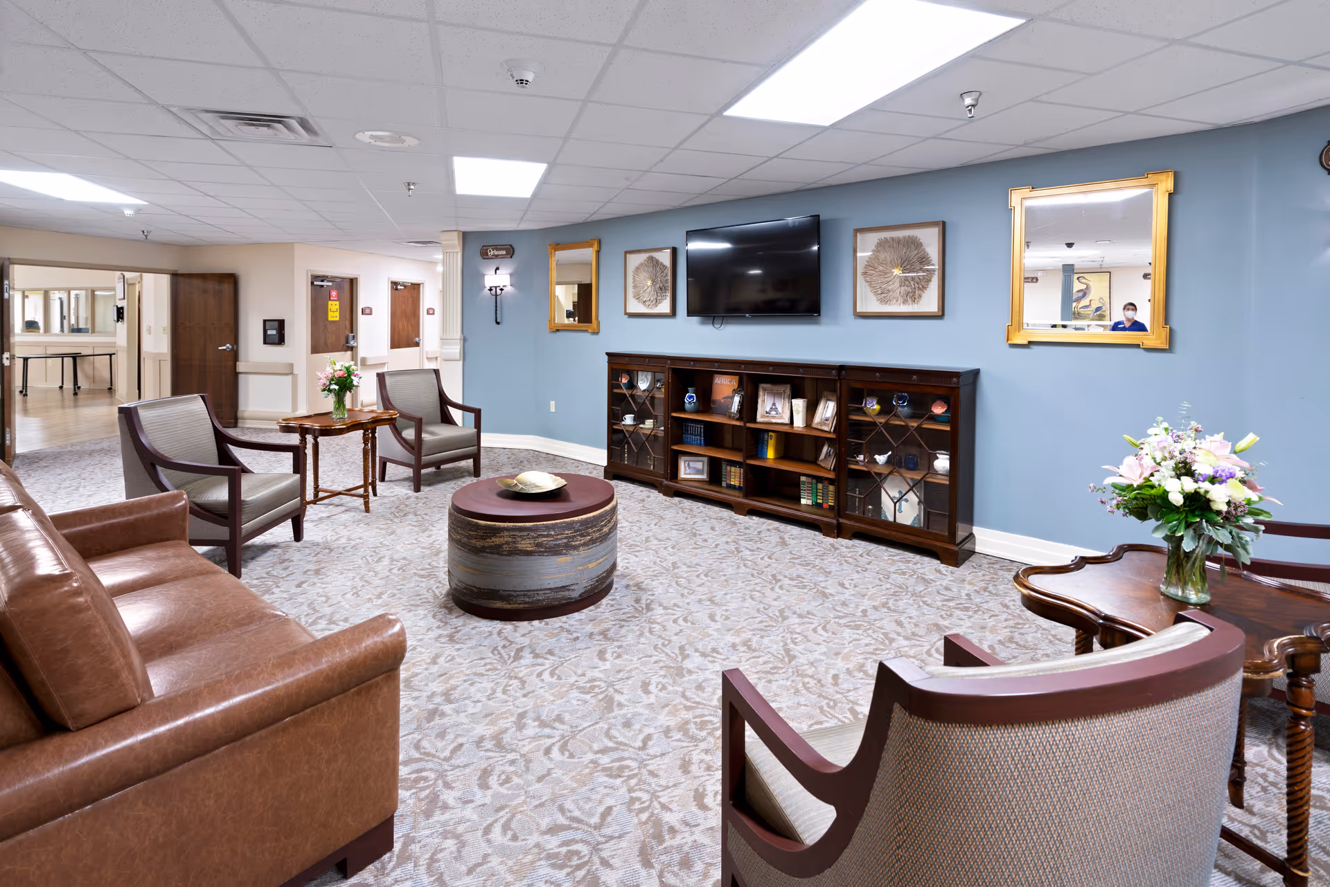 A spacious senior living common room with leather sofas and armchairs arranged around a round ottoman, a TV and bookshelf on a blue wall, and floral arrangements on side tables.