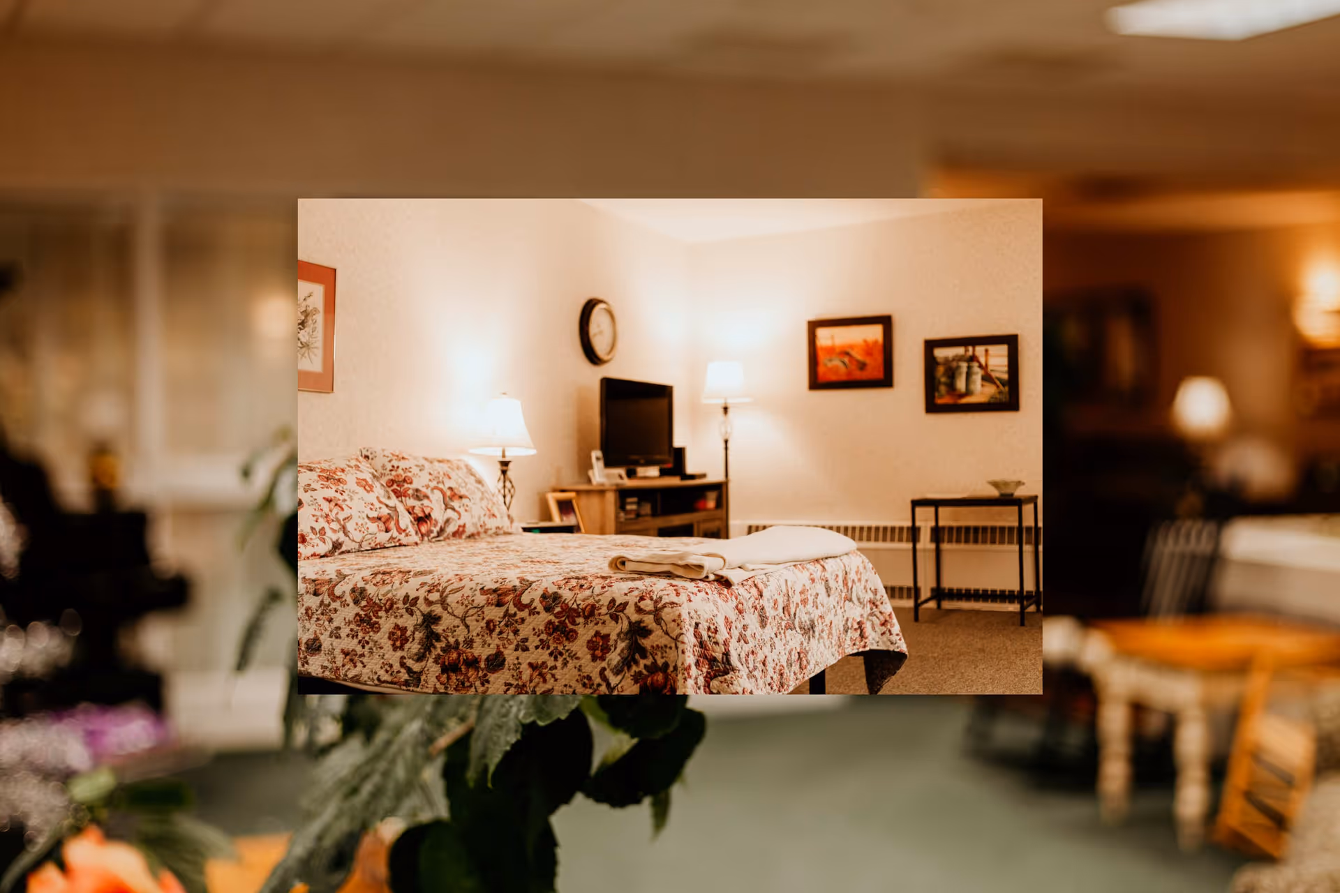 A cozy bedroom with a floral bedspread, bedside lamps, a TV on a dresser, and framed pictures on the wall.