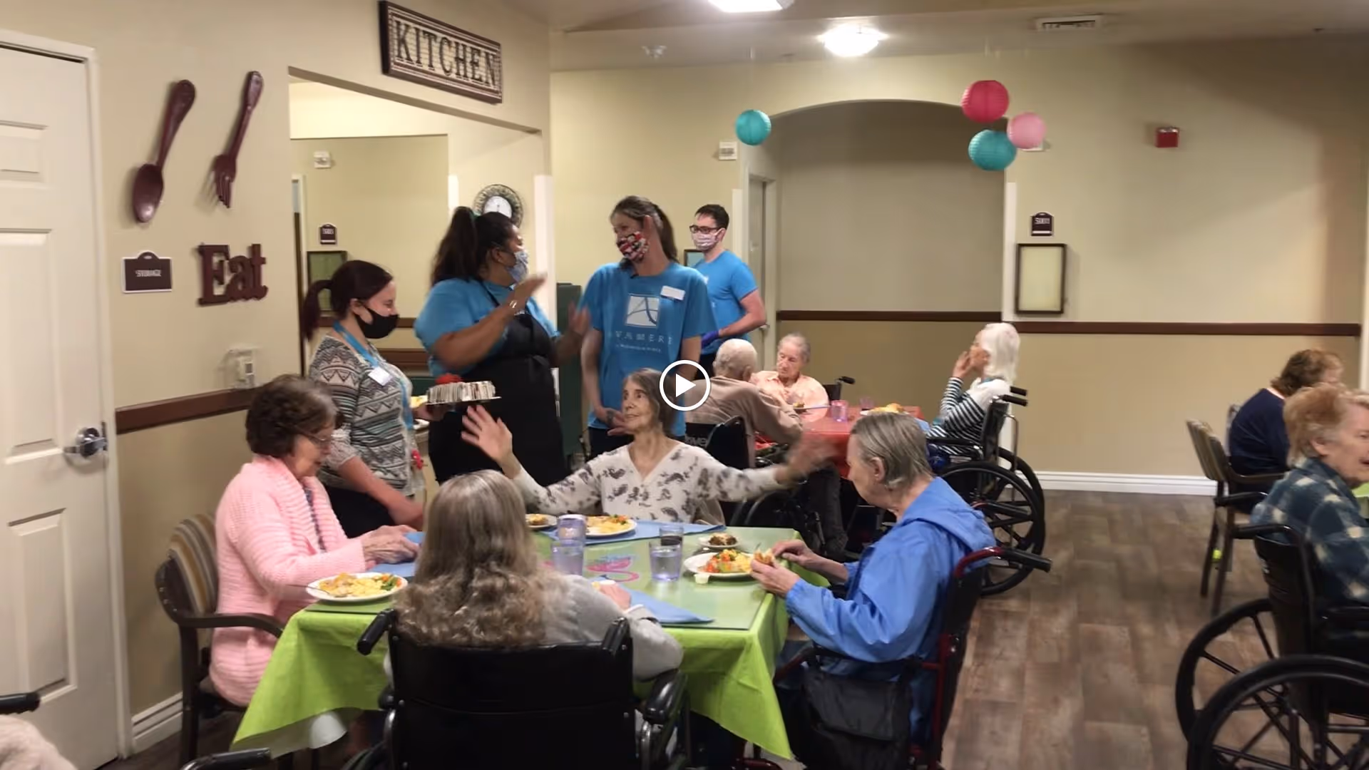 A group of elderly residents seated at tables covered with green tablecloths, eating meals in a dining area. Several staff members wearing masks and blue Avamere shirts are interacting with the residents. The room has beige walls with decorations including large wooden utensils and the word 'Eat' on the wall near a door labeled 'Storage'. Colorful paper lanterns hang from the ceiling.