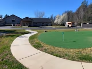 A curved concrete pathway leading around a small putting green with a flag, situated in front of a single-story building with a brown roof. Trees and shrubs are visible in the background under a clear sky.