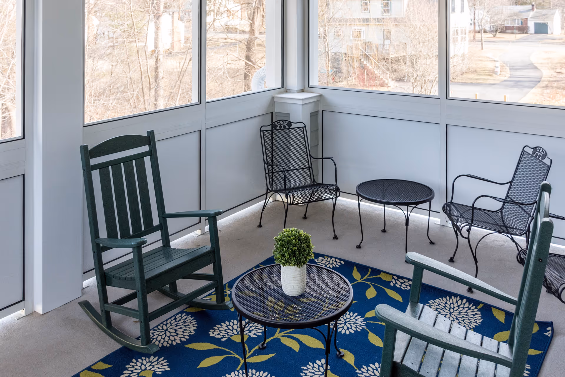 A screened-in porch area with two green wooden rocking chairs, two black metal mesh chairs, and two black metal mesh tables. A blue rug with white and green floral patterns is on the floor, and a small potted plant is on one of the tables. Outside the porch, leafless trees and houses are visible.