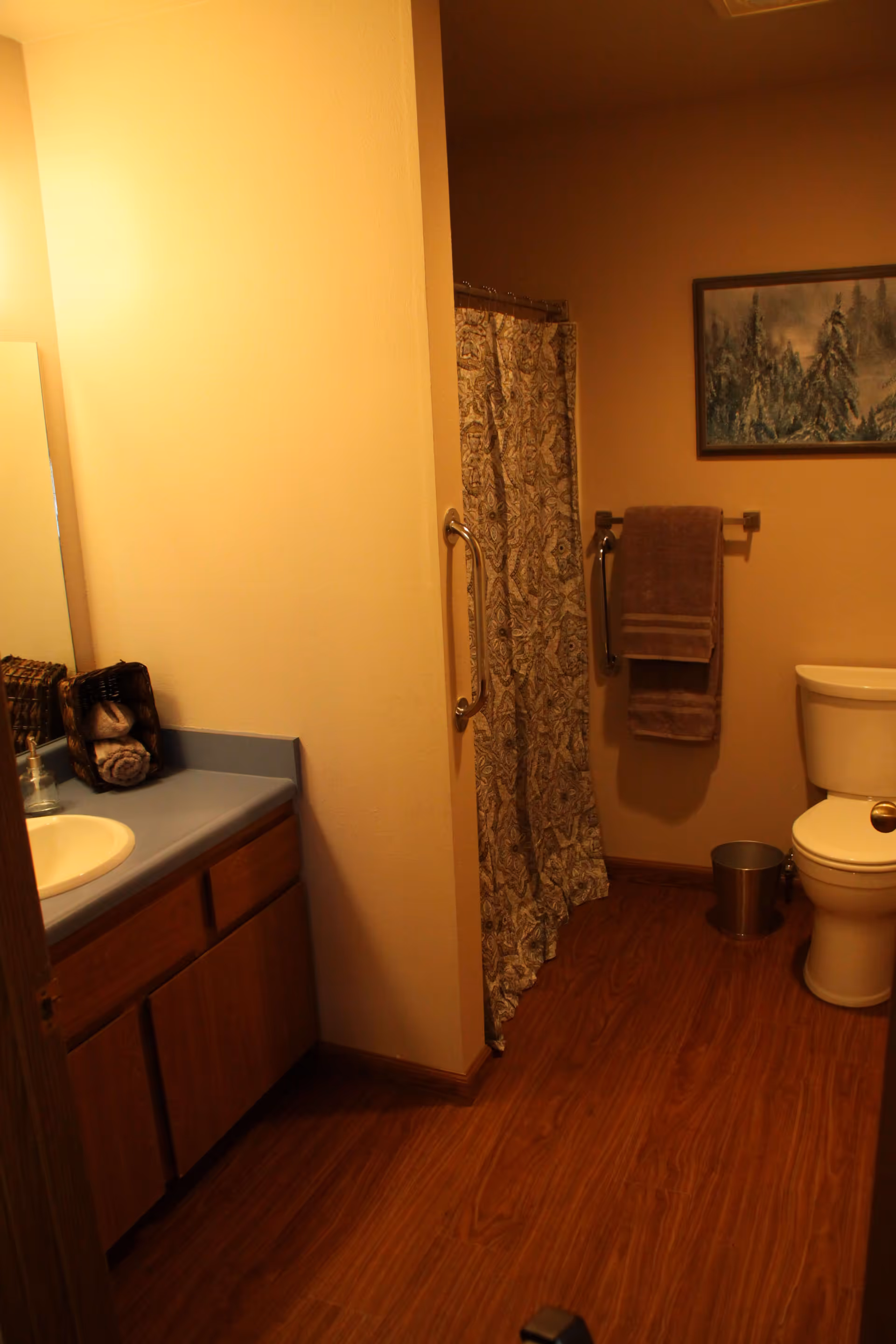 A bathroom with wooden flooring, a white toilet, a towel rack with brown towels, a shower with a patterned curtain, a sink with a blue countertop, a wicker basket with rolled towels, and a framed painting of a snowy forest on the wall.