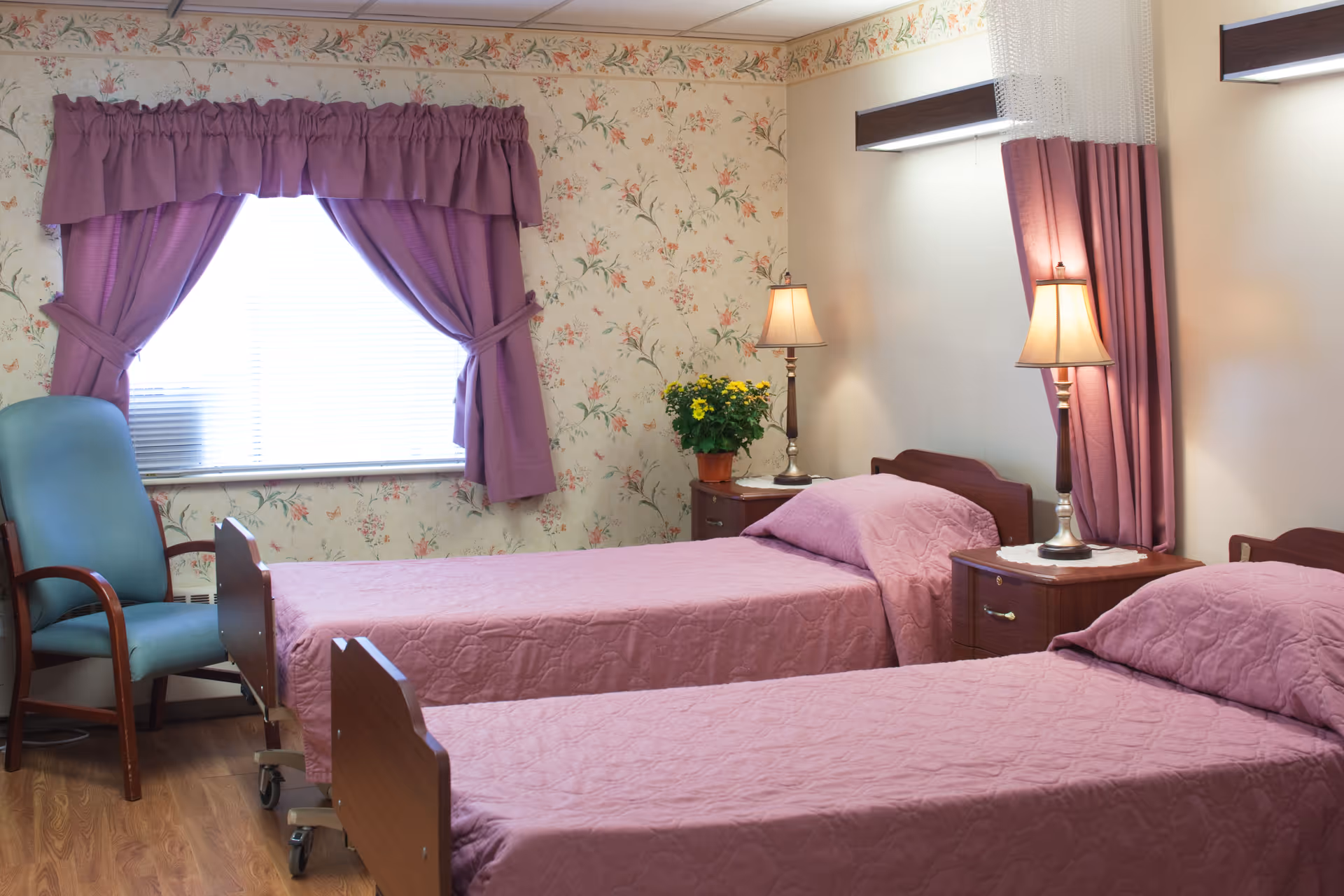 A cozy bedroom with two single beds covered in matching purple bedspreads. Each bed has a wooden headboard and is accompanied by a wooden nightstand with a lamp. There is a window with purple curtains and a floral valance, a green upholstered chair, and a potted yellow flower on one nightstand. The walls have floral wallpaper and the floor is wooden.