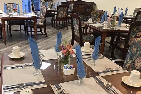 Dining room with multiple wooden tables set with placemats, silverware, cups, and glasses with blue folded napkins. Floral upholstered chairs surround the tables, and a small flower arrangement is placed in the center of each table.