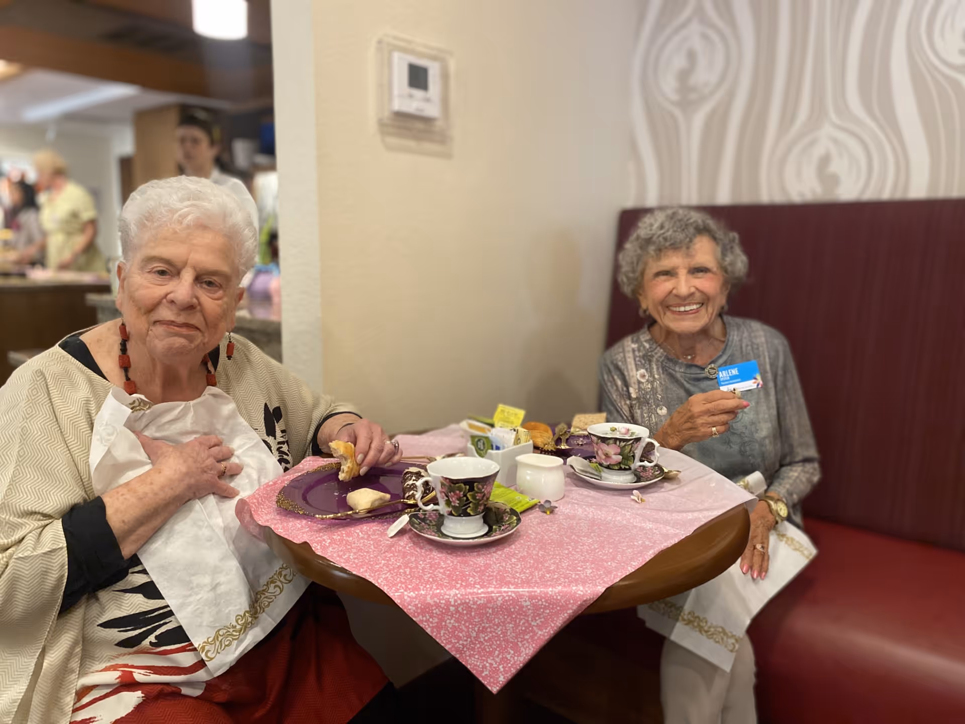 Two elderly women sitting at a small round table with a pink tablecloth, enjoying tea and snacks. One woman is holding a piece of food and wearing a white napkin, while the other woman is smiling and holding a name tag that reads 'Arlene'. The background shows a cozy indoor setting with patterned wallpaper and a maroon cushioned bench.