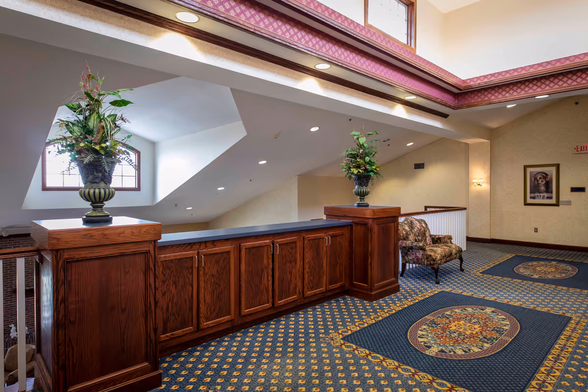 Interior view of a senior living facility hallway with wooden cabinetry topped with decorative flower arrangements, patterned carpet with floral designs, a floral upholstered armchair, and a framed picture on the wall. The ceiling has recessed lighting and a skylight with patterned trim.