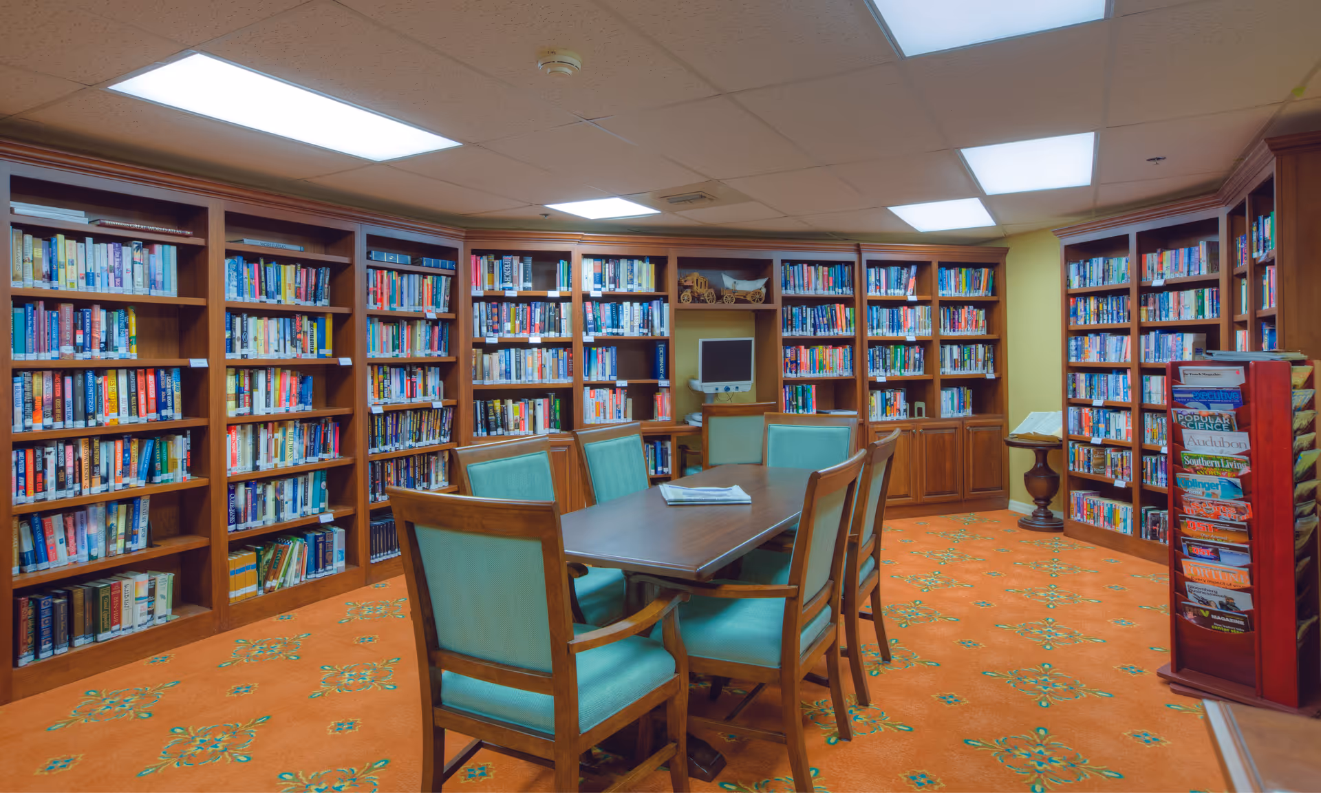 Cozy library room with wooden bookshelves lining the walls, a central table surrounded by green-upholstered chairs, and a magazine rack.