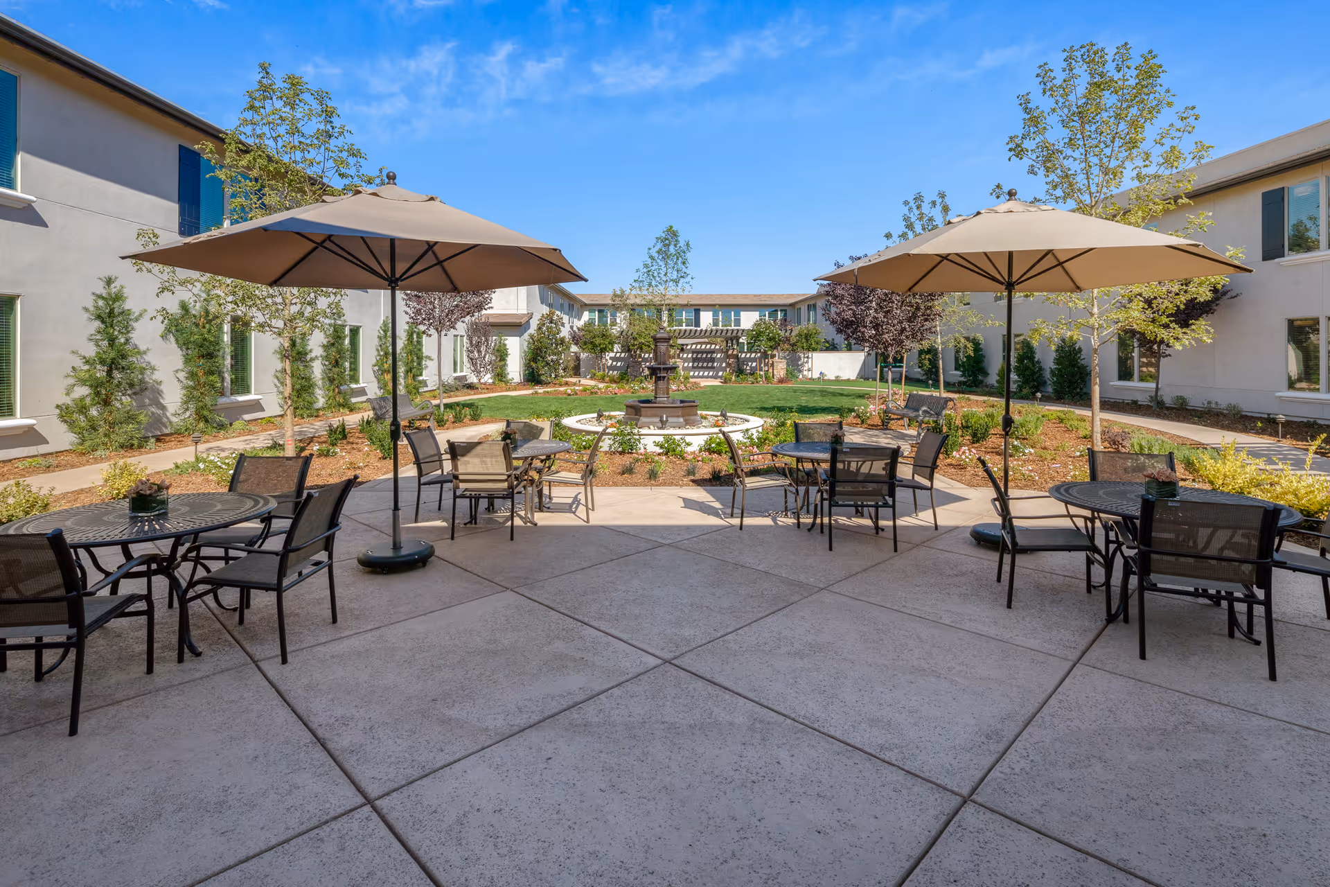Outdoor courtyard area at Oakmont of El Dorado Hills featuring round tables with chairs and large beige umbrellas on a concrete patio, surrounded by landscaped garden beds with trees and shrubs, and a central water fountain with a grassy area and buildings in the background under a clear blue sky.