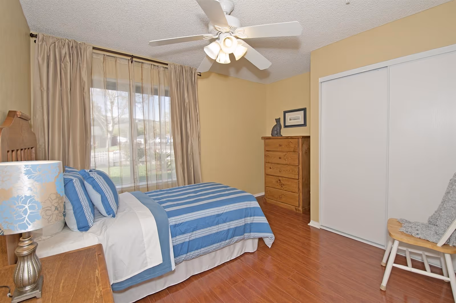 Bright bedroom with a single bed dressed in blue striped linens, wooden dresser, ceiling fan, large curtained window and sliding closet doors.