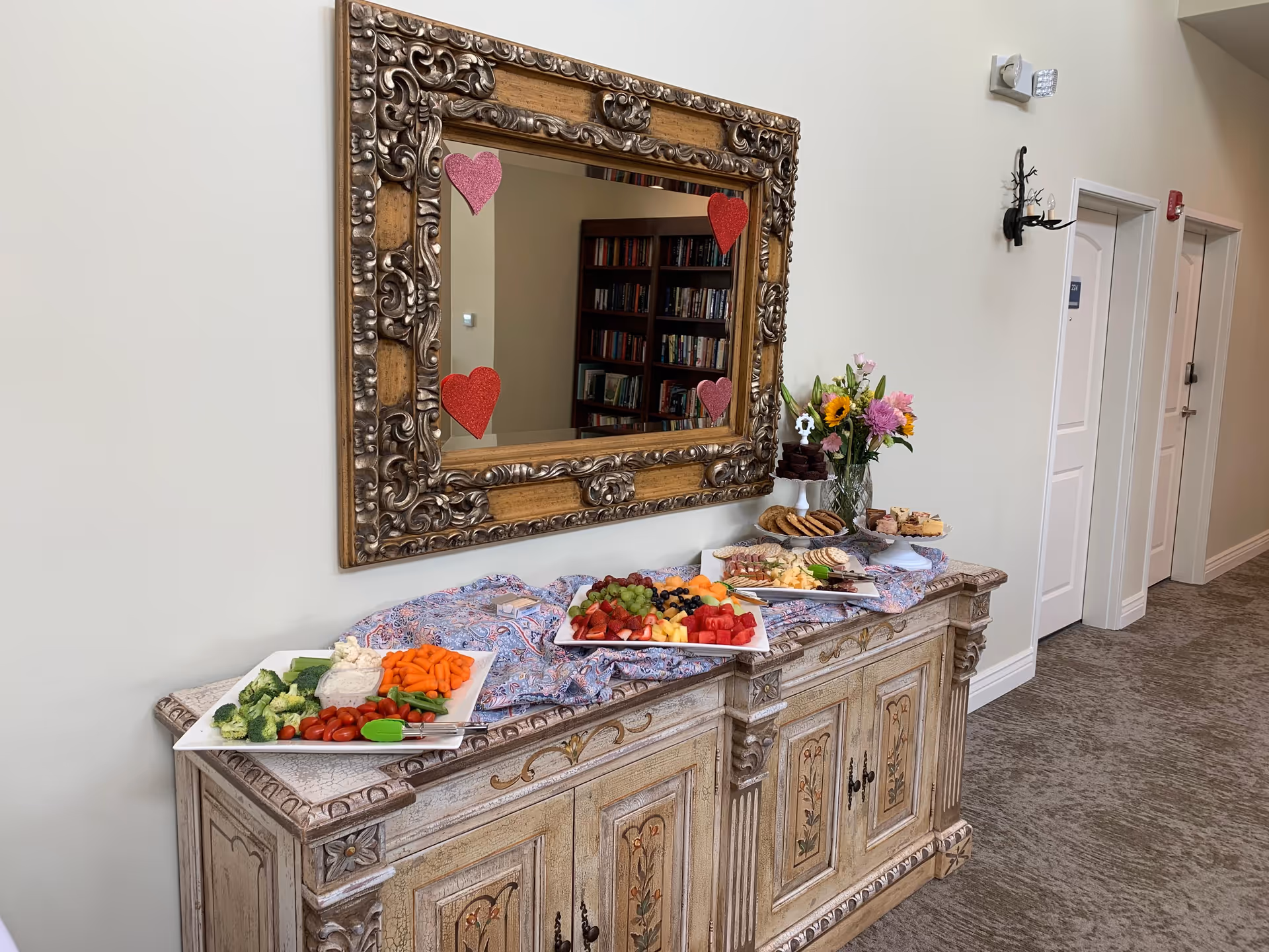 A decorative wooden sideboard topped with a patterned cloth and an assortment of food platters including fresh vegetables, fruit, cookies, and desserts. Above the sideboard is an ornate framed mirror with red and pink heart decorations. A vase with colorful flowers is also on the sideboard. The setting appears to be a hallway with closed white doors and a carpeted floor.