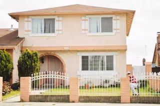 Two-story beige house with white trim, arched entry, metal fence, and a small lawn.