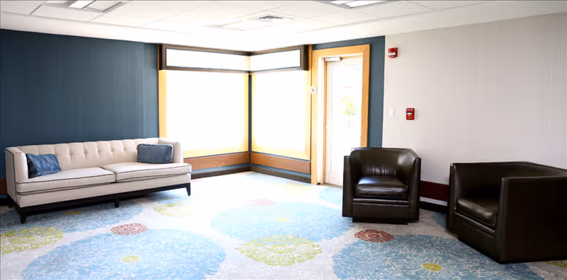 A bright and spacious living room area with a beige tufted sofa with two blue cushions on the left, two dark brown leather armchairs on the right, large windows with natural light, and a colorful carpet with blue, green, and red circular patterns.