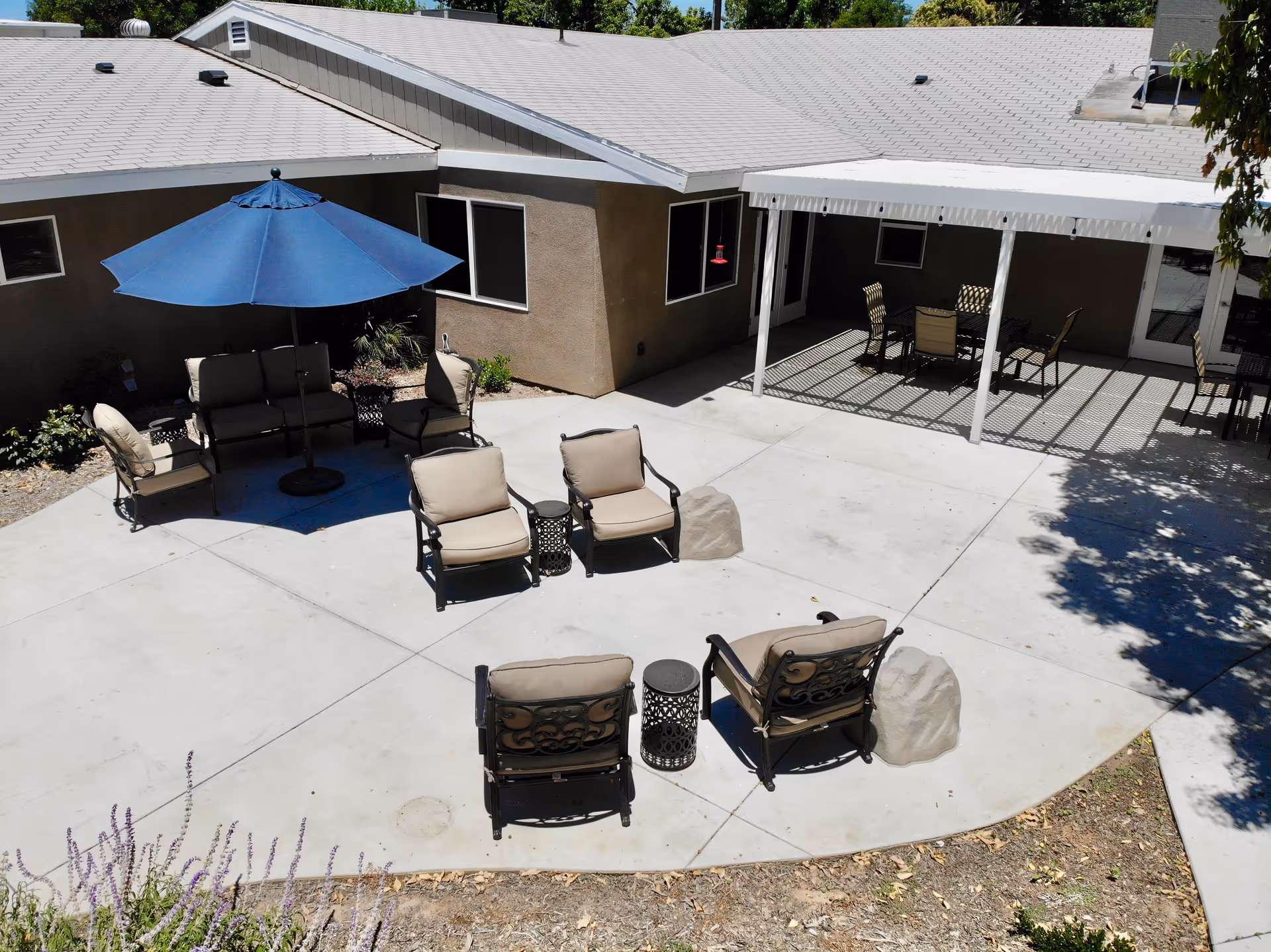 Outdoor patio area at The Hills of Stillman featuring cushioned chairs arranged around small tables, a blue umbrella providing shade over a seating area, and a covered section with additional chairs and tables. The patio is surrounded by a building with beige walls and windows.