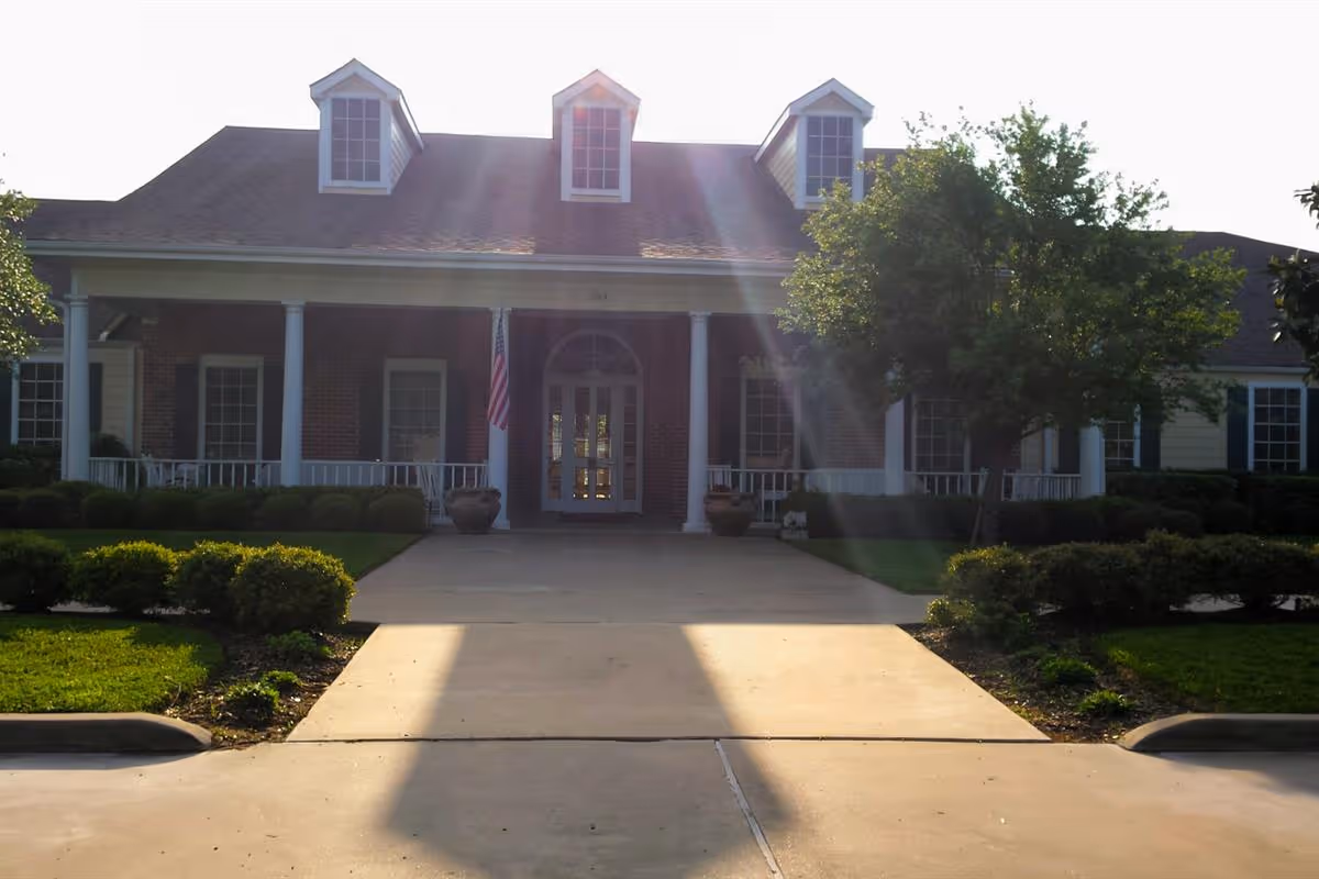 Front exterior view of a single-story brick building with white columns, a porch, and three dormer windows on the roof. There is a concrete driveway leading up to the entrance, with neatly trimmed bushes and a tree on the right side. Sunlight is shining from behind the building.