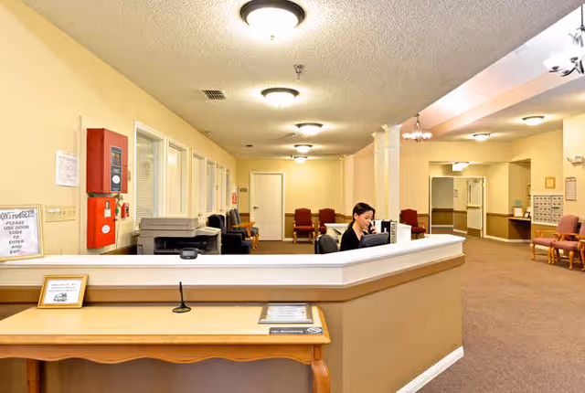 Reception area inside Sabine Place facility with a woman seated behind a large white reception desk, multiple chairs along the walls, beige walls and carpet, ceiling lights, and a fire alarm box on the left wall.