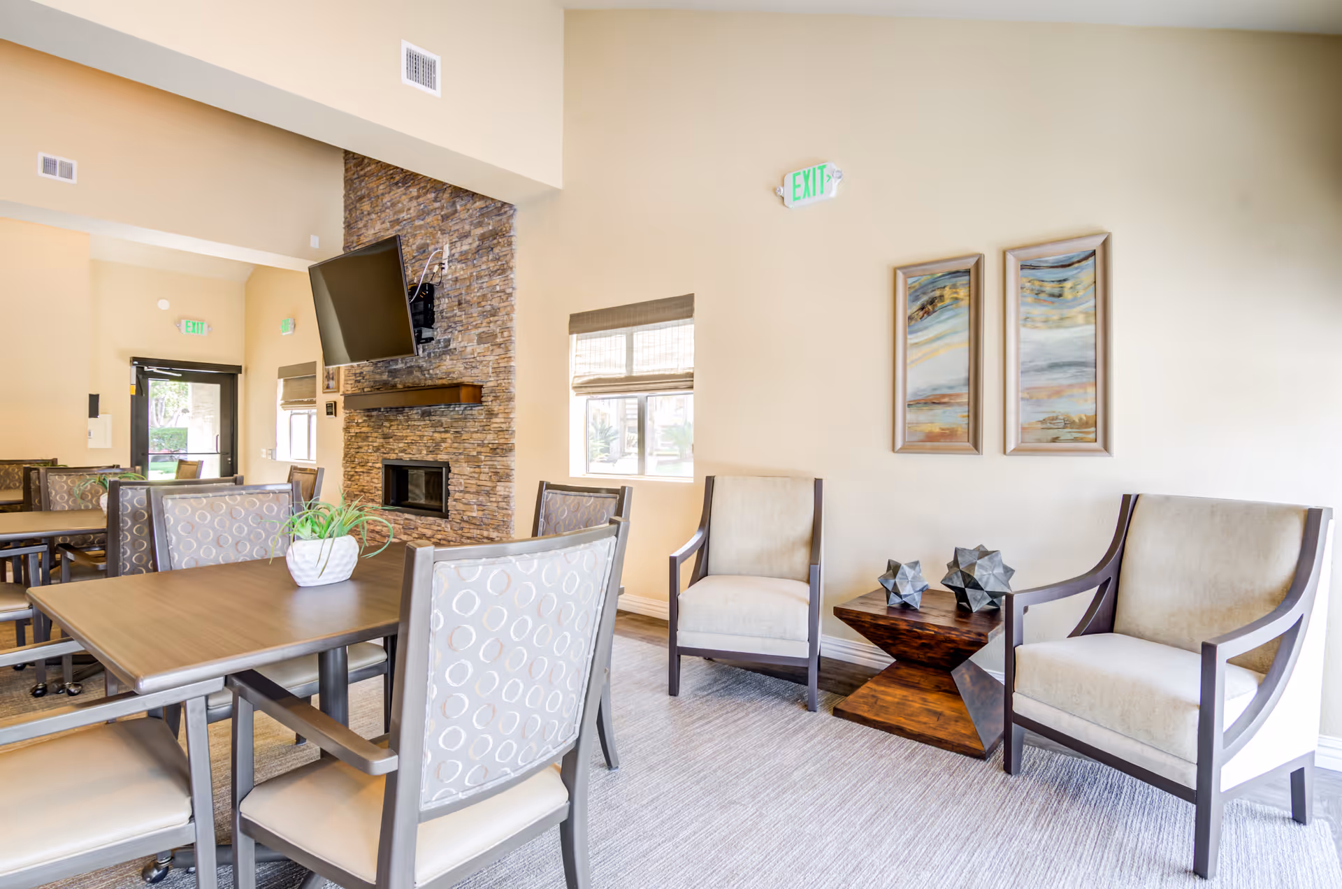 A bright senior living facility common area with beige walls, a stone fireplace with a mounted flat-screen TV, several tables and chairs with patterned upholstery, two beige armchairs flanking a wooden side table with decorative objects, and two abstract paintings on the wall.